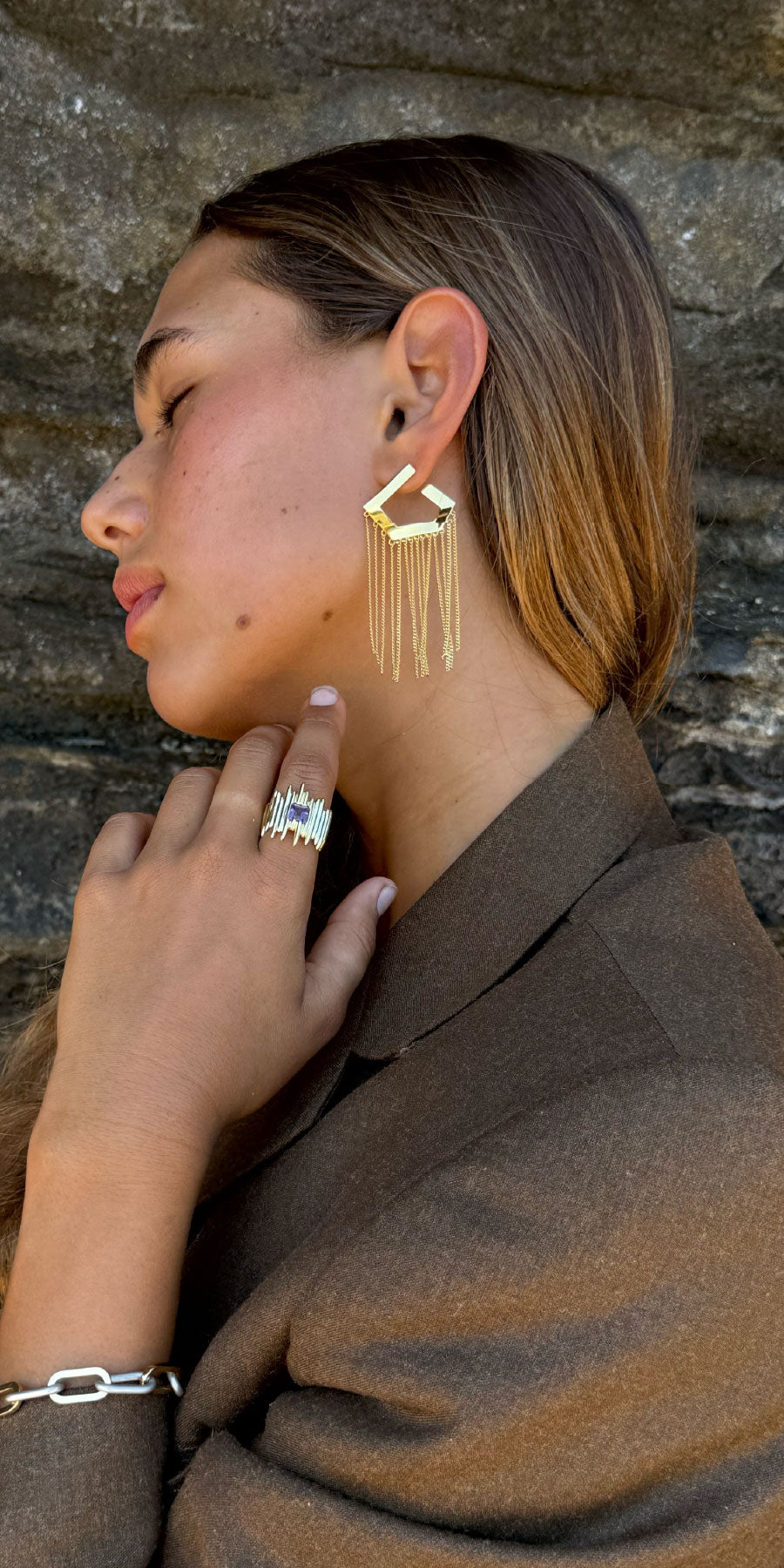 Woman wearing gold tassel earrings and a ring, against a stone wall background